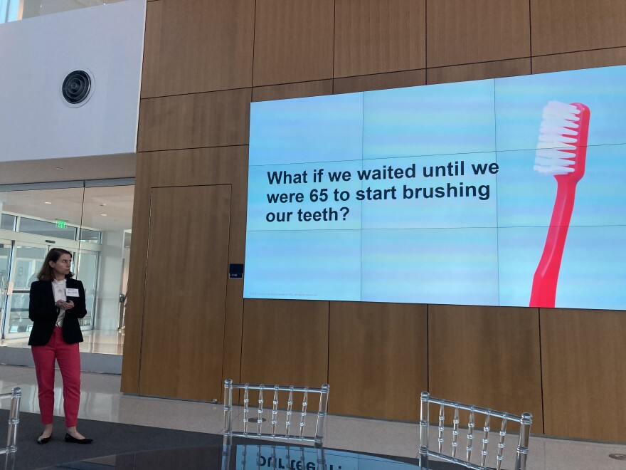 A person stands in front of a slide with a large cartoon toothbrush, reading," What if we waited until we were 65 to start brushing our teeth?"