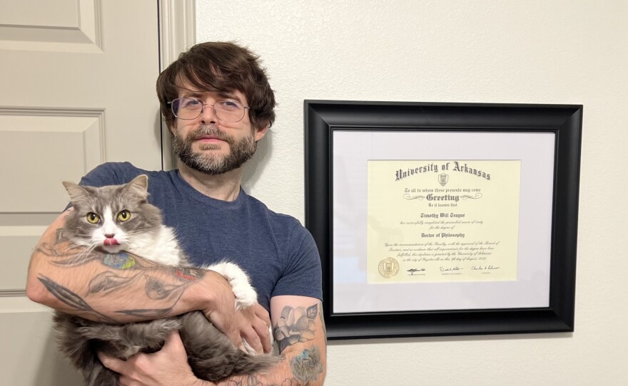 A man with shaggy brown hair, a brown beard, glasses and multiple tattoos on his arms stands by a diploma hanging on the wall while holding a cat.