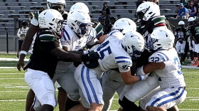 football action showing several players in a white uniform with blue strips tackling a player in a black jersey. both teams wearing white helmets. set on a green field with white football lines