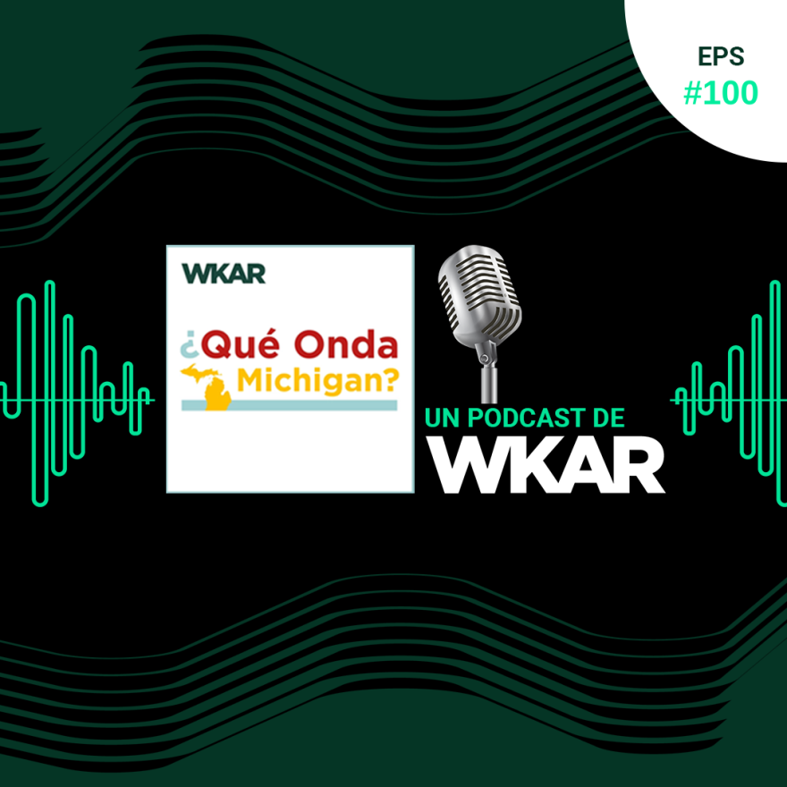 La imagen muestra un micrófono en un fondo negro. El micrófono es de color plateado y tiene una forma cilíndrica. Hay una rejilla en la parte delantera del micrófono para proteger la cápsula del micrófono. El micrófono está apoyado sobre un soporte de metal. En la parte superior del soporte hay una etiqueta que dice "EPS #100 WKAR ¿Qué Onda Michigan? UN PODCAST DE WKAR".