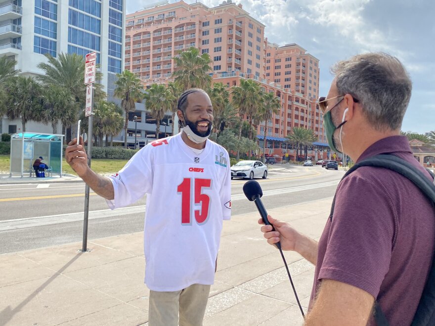 A reporter wearing a mask and holding a mic interviews a man wearing a chiefs jersey with his mask under his chin.