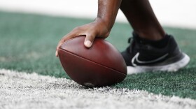A close up shot of an unidentifiable person off screen grabbing a football. The brown oval shaped ball is in the center of the image, pressed against the ground by a hand whose arm cuts off right above the wrist. The person's foot can also be seen in the image, just behind the ball. Their wearing a black Nike tennis shoe with the white outline of Nike's check mark symbol imprinted on the side of the shoe. 