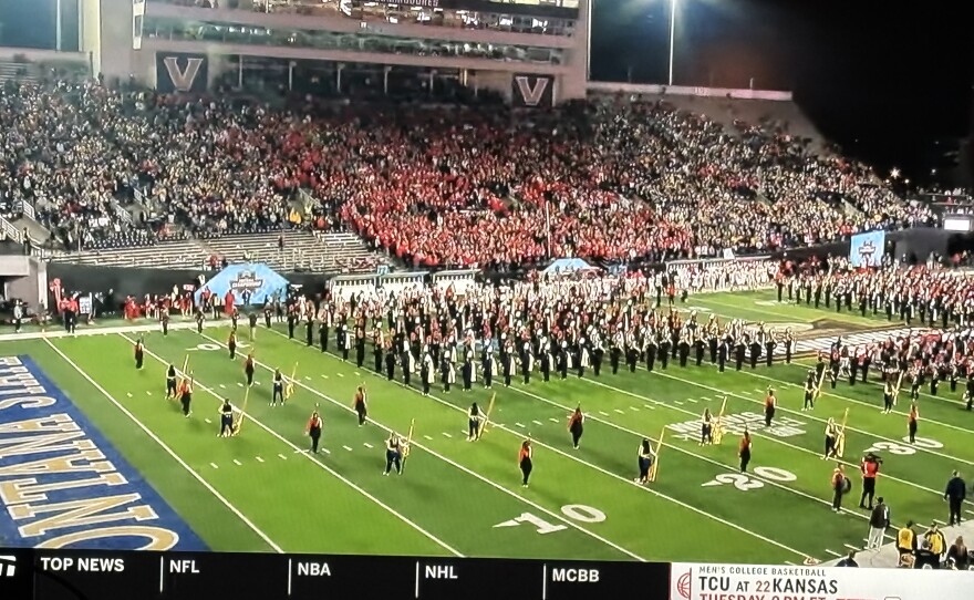 A college football stadium with a large marching band performing on the field. The stands are full of fans, many wearing red, and stadium lights illuminate the scene at night. A TV sports ticker runs along the bottom of the screen.