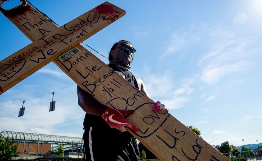 A man carrying a wooden cross also wears a mask, gloves and glasses during a march near Music Hall as people continue to rally and protest the murder of George Floyd as the coronavirus pandemic continues, Wednesday, June 3, 2020, in Cincinnati.