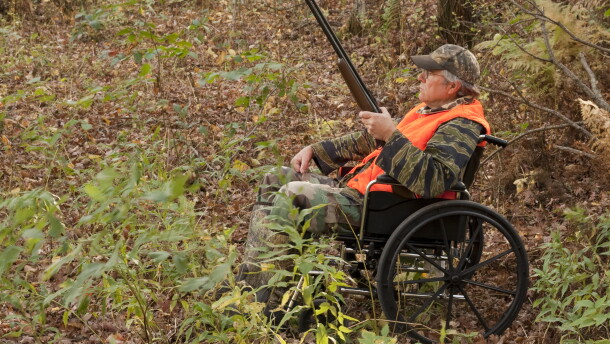 A hunter using a folding-frame wheelchair sits in a wooded area during fall. He is holding a shotgun and wearing camouflage clothing with a bright orange safety vest and camouflage cap.