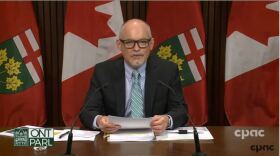 Dr. Kieran Moore sits at a brown desk, reading from a white paper, in front of two microphones.