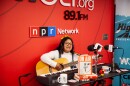 Samara Jane, winner of the 2025 Highway 309 Studio Sessions Contest, performs at WGLT. She sits in front of a red WGLT and NPR Network backdrop, playing an acoustic guitar and singing into a studio microphone, with a keyboard and radio gear on the table in front of her.