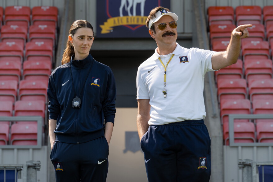 A woman and man in soccer team apparel standing in front of empty stadium seats as they look out at a soccer pitch.