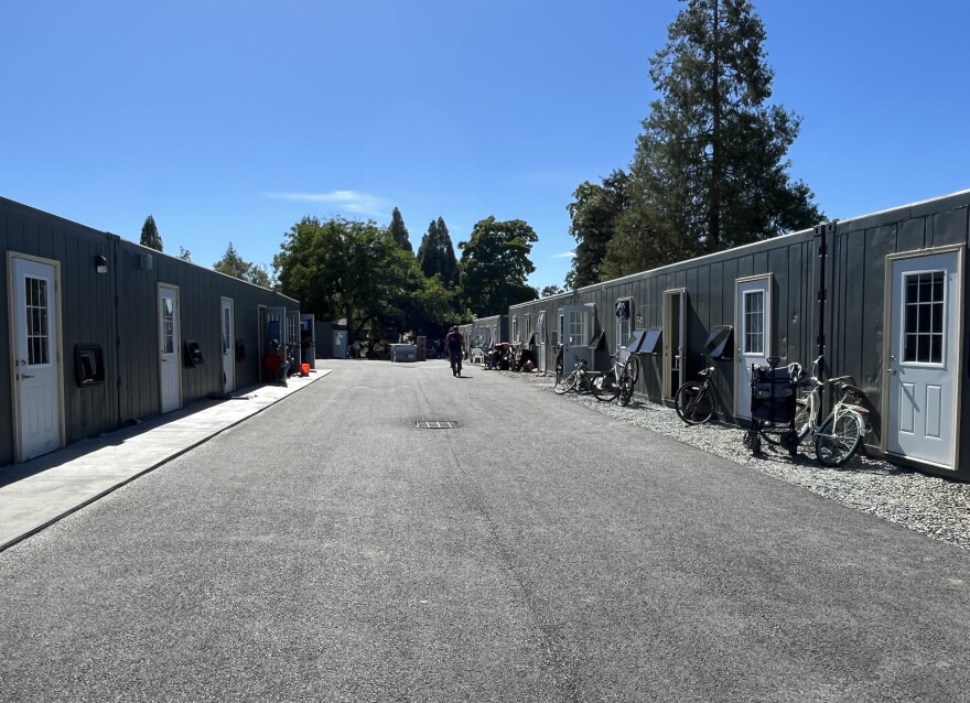 A wide driveway is lined with single-story shipping container homes. They are gray and have white doors and windows. 