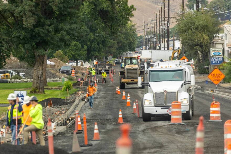 A large white truck surrounded by orange traffic cones and men in safety vests and hard hats in the foreground show construction underway on Snake River Avenue in Lewiston. 