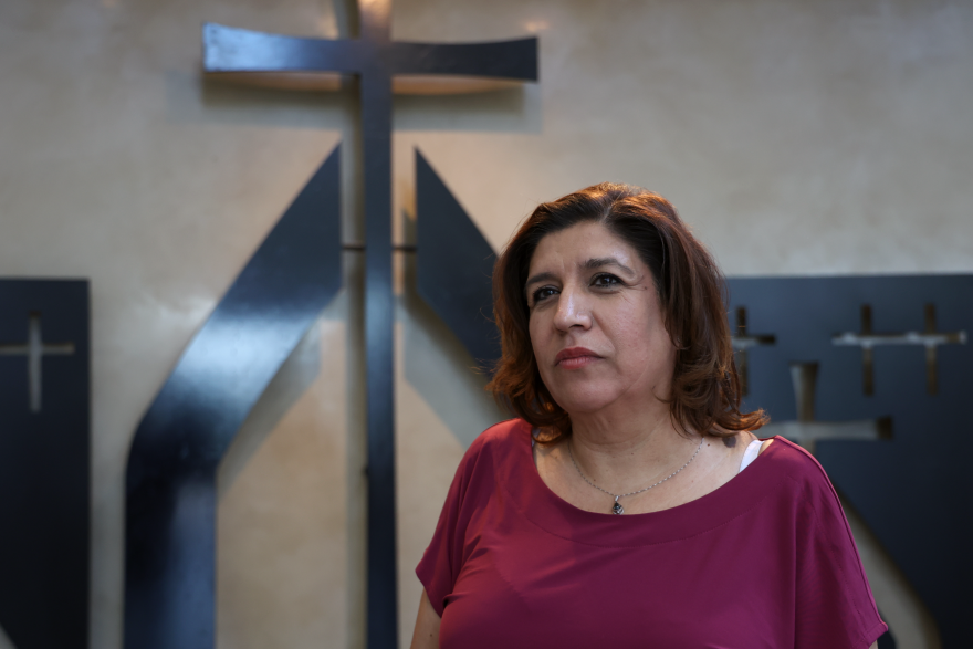 A woman in a maroon shirt stands in front of a cross.