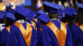The backs of a crowd of graduates in blue graduation gowns wearing gold sashes