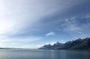 A large blue lake with sparse clouds in a blue sky, with snow-peaked mountains on one shore and smaller mountains in the far distance.