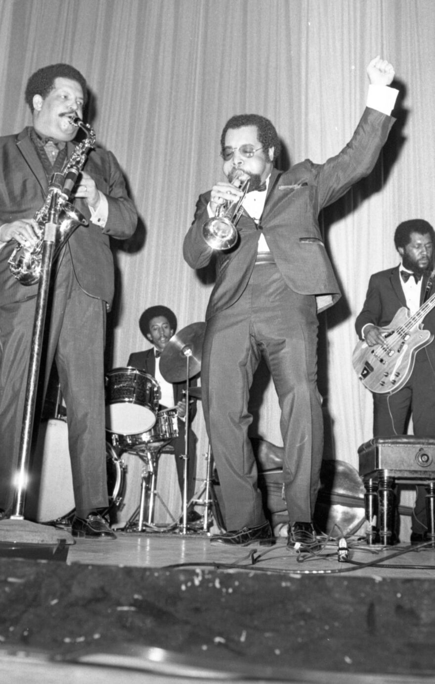 Two Black men in suits, on stage. One plays a saxaphone while the other plays a trumpet