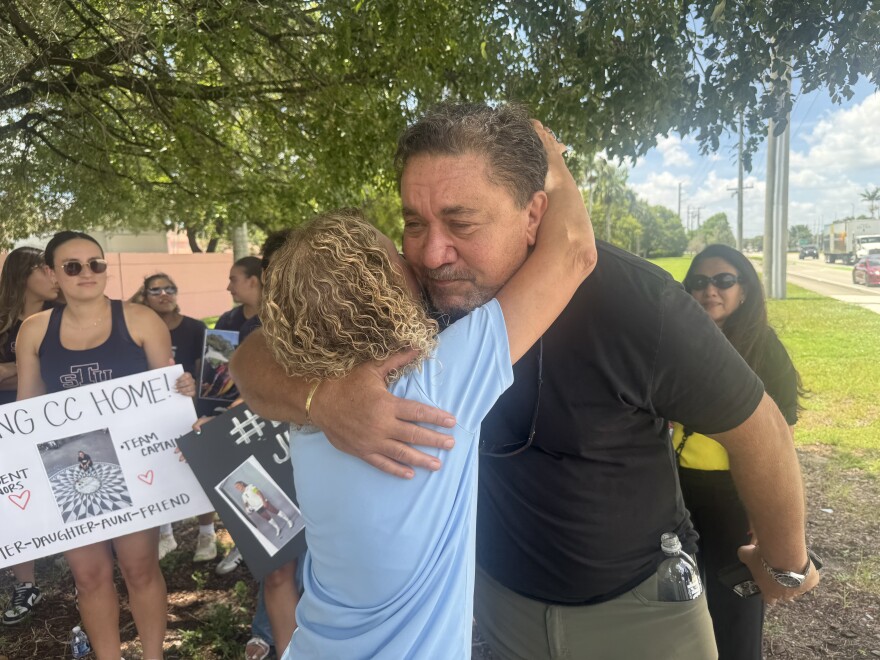 A shorter blonde woman in a blue top hugs a taller man in a black shirt