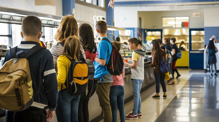Students standing in line at a school cafeteria, waiting to get their lunch. They carry backpacks and engage in casual conversation.