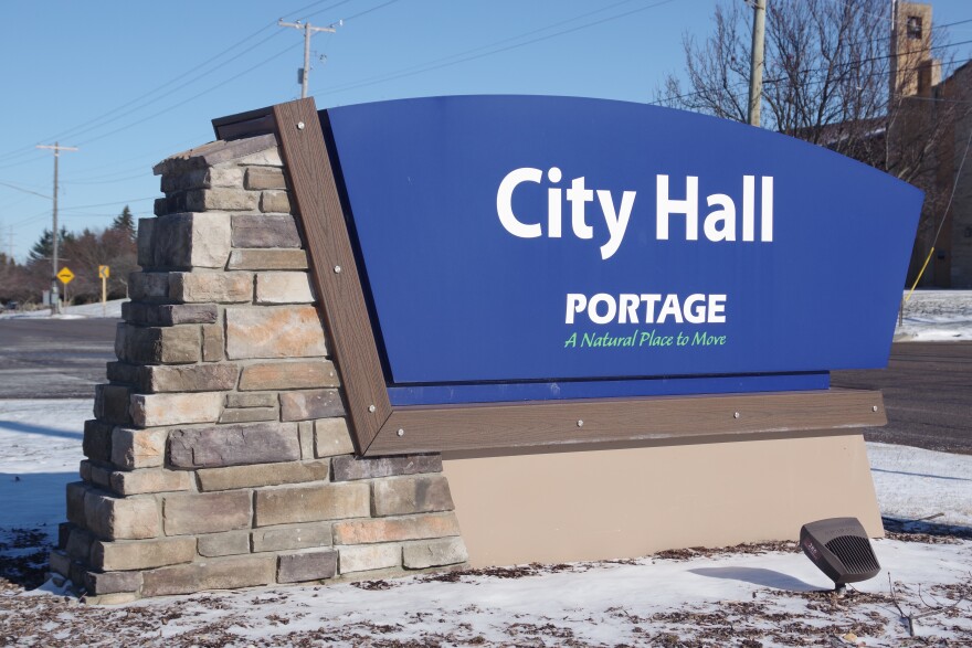 A large sign marks the location of Portage City Hall. On the left side of the sign, stakes of rectangular cut stones build up to the top. A majority of the middle and right of the sign is taken up by a large blue segment that reads "City Hall," with "Portage" and its motto "A Natural Place to Move" sitting below the sign in that order. 