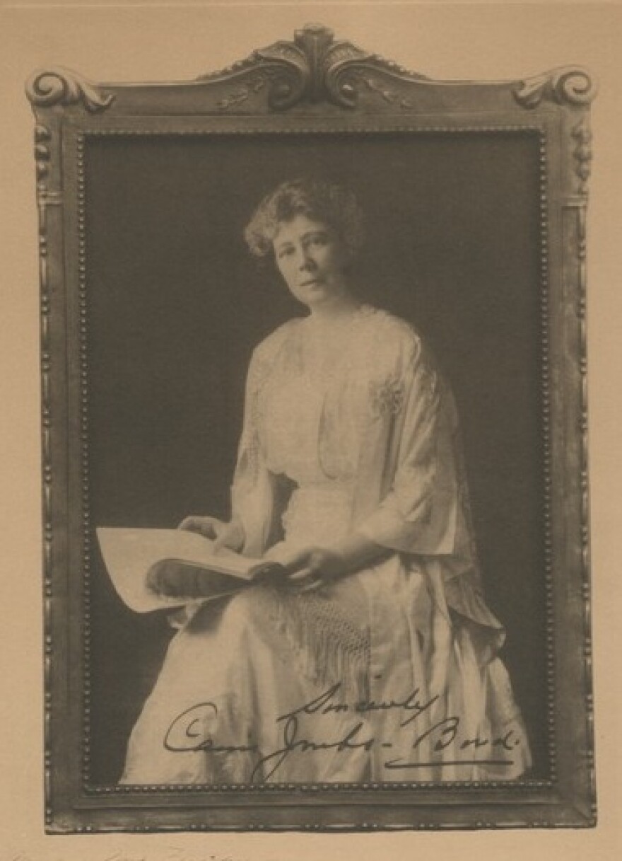 Carrie Jacobs-Bond is seated in a white dress holding sheet music. 