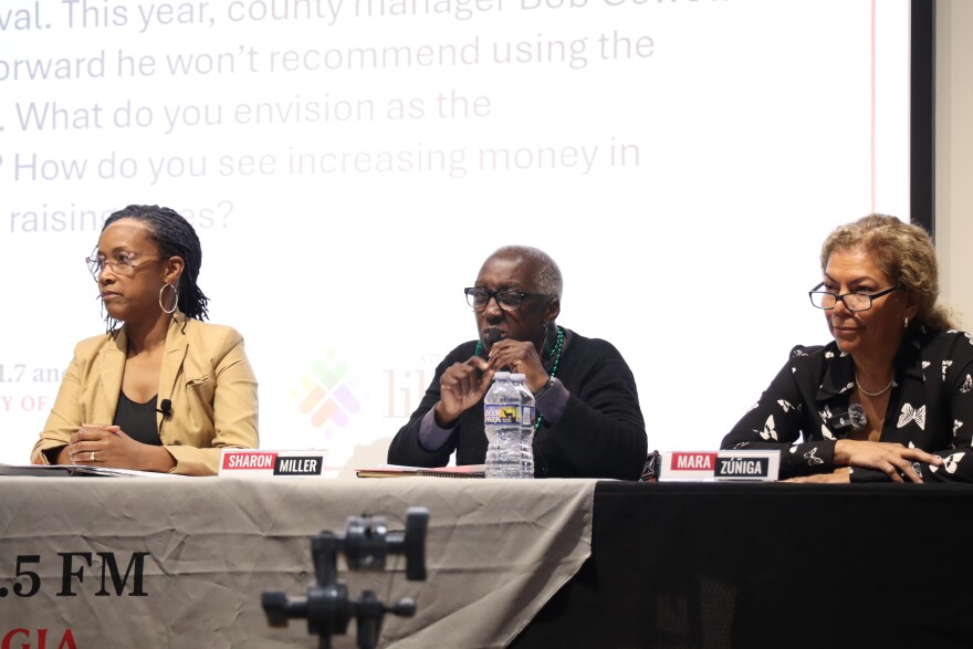 Three candidates sit at a table during an Athens mayoral debate, with one speaking into a microphone.