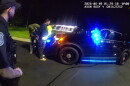 Two men are being handcuffed by a border patrol agent as they stand in front of the hood of a police car. In the foreground, a police officer with a Southgate police emblem on his shoulder watches the scene. It is dark, but a brightly-lit gas station can be partly seen in the background. 