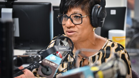 A woman sits inside a radio studio wearing a headset and talking while gesturing with her right hand.