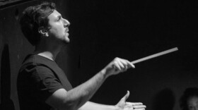 Matt Marco, Artistic Director for Buffalo Opera Unlimited, is holding a baton as he conducts an orchestra.