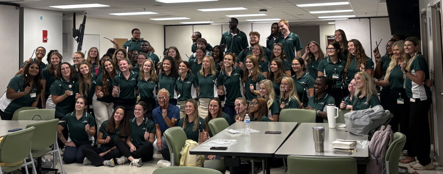 A group of college students wearing green shirts hold up magic wands with magician Kevin Spencer in the middle.