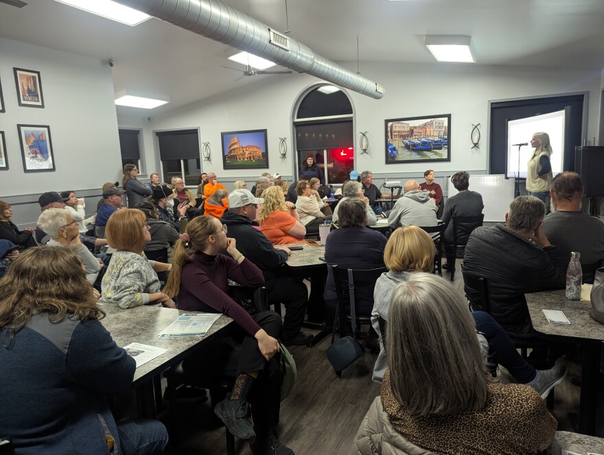 A crowd of people sit at tables to listen to a presentation.