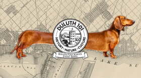 A dachshund dog, stretched out over an old map of a town; a black and white emblem in front with "Duluth 101" written on it