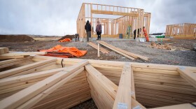 A bare structure built with wooden two-by-fours has several people standing around it. There is wood in the foreground of the picture.