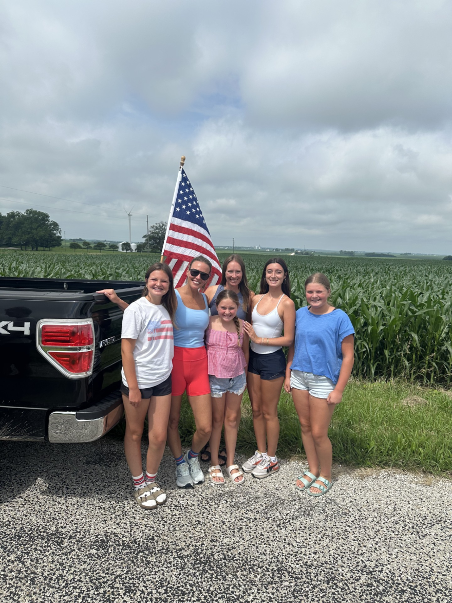 Nicole Roberts stands with five other girls and an American flag outside in front of a corn field. 