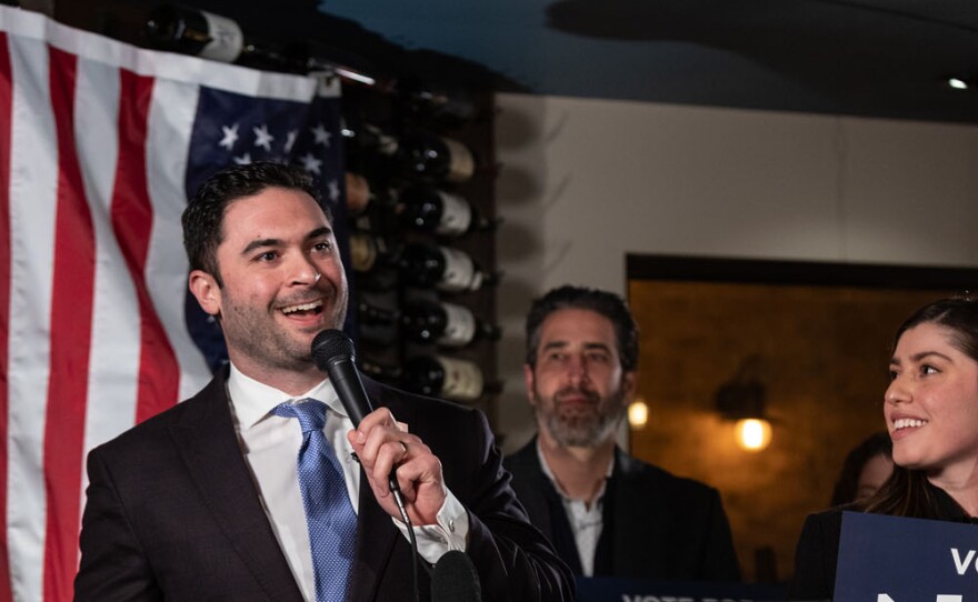 A man with short dark hair is standing behind a podium and speaking into a micophone.