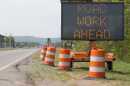 A highway sign saying "road work ahead" sits alongside four large orange and white traffic construction pylons.