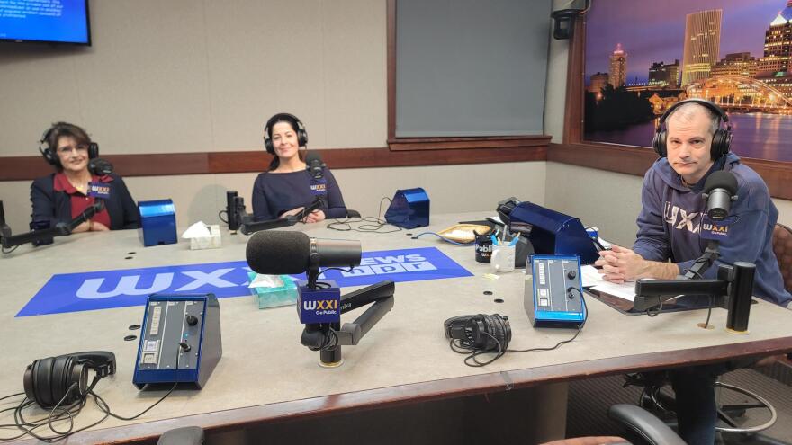 Three people wearing headphones sit at a table in a radio talk studio: a woman at left has brown hair and is wearing glasses, a navy blazer and a red button-down shirt; a woman at center has her dark hair pulled back in a ponytail and is wearing a navy top; a man at right has very short dark hair and is wearing a navy hooded sweatshirt with white lettering.