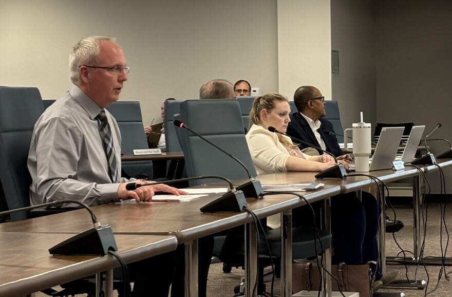 A man in business attire sits at a table, and speaks into a microphone.  Three other people sit at the table and look to the left, at a screen listing budget information.