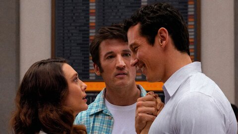 A still from the film Eternity. Elizabeth Olsen’s Joan and Callum Turner’s Luke stand close, hands clasped while Miles Teller’s Larry looks on. 