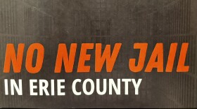 An image of a small postcard-like document that reads in red and white text "NO NEW JAIL IN ERIE COUNTY". In the background is a darkened image of inside a jail cell.