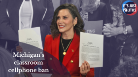 Michigan Gov. Gretchen Whitmer, wearing a red blazer, holds up two signed bills during a ceremonial signing. Text on the image reads, “Michigan’s classroom cellphone ban,” with the “It’s Just Politics” logo in the top corner.