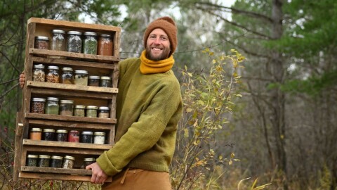 A man stands outdoors holding a wooden shelf filled with glass jars of dried herbs and other ingredients.
