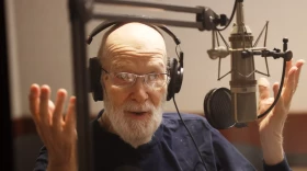 balding man with grey beard with headphones at microphone, gesturing with hands