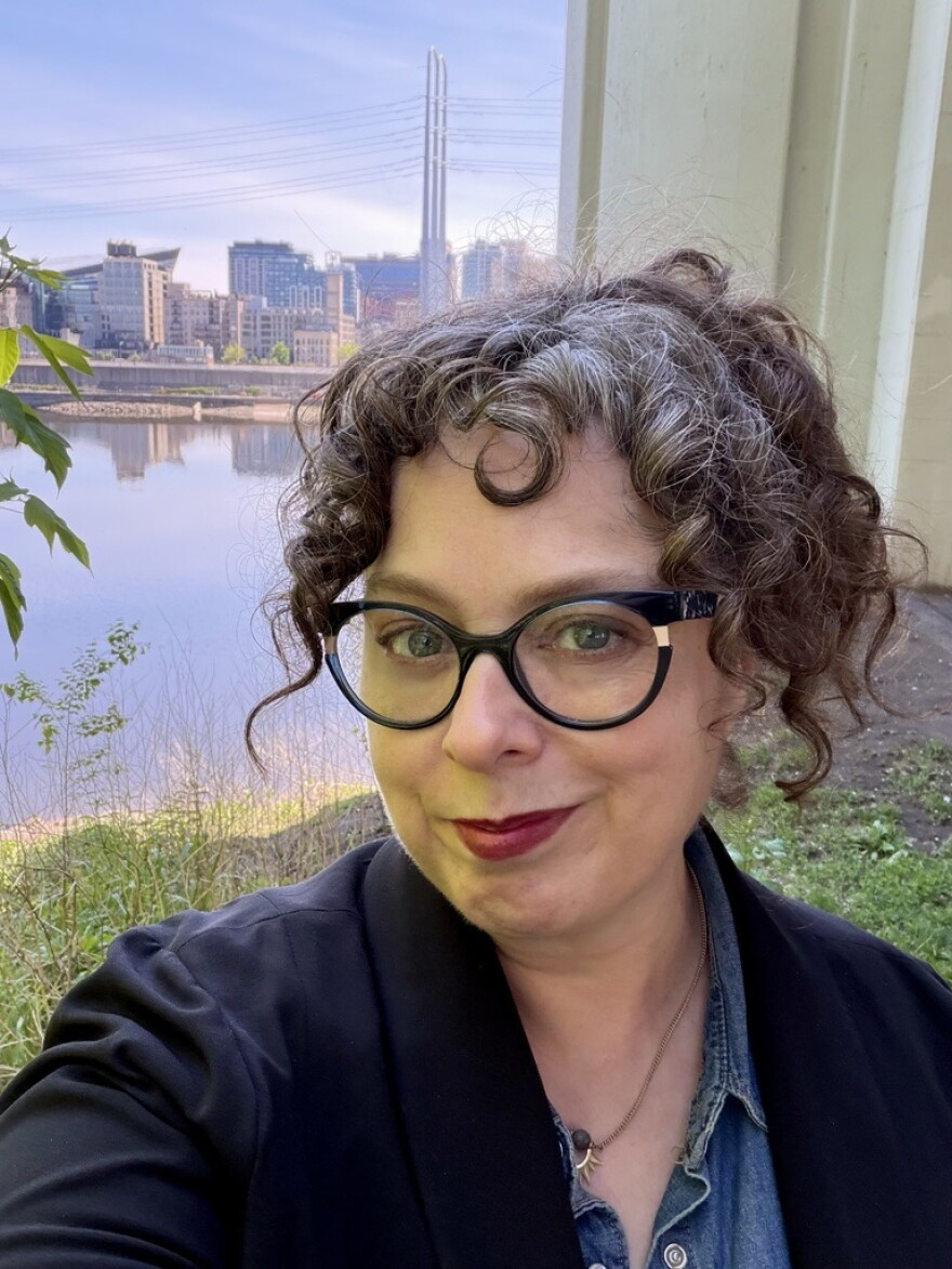 A woman with brown curly hair and glasses poses in front of a river in a city. 