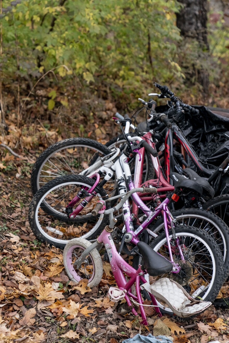 Children’s bikes are stacked in a yard. The smallest, a pink child's bicycle, is missing a back wheel. 