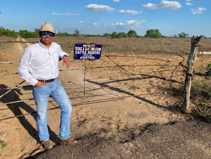 Whit Jones wears jeans, a white button-down shirt, sunglasses and a straw cowboy hat. He leans against a beat-up cattle fence, which bears the Texas & Southwestern Cattle Raisers Association sign.