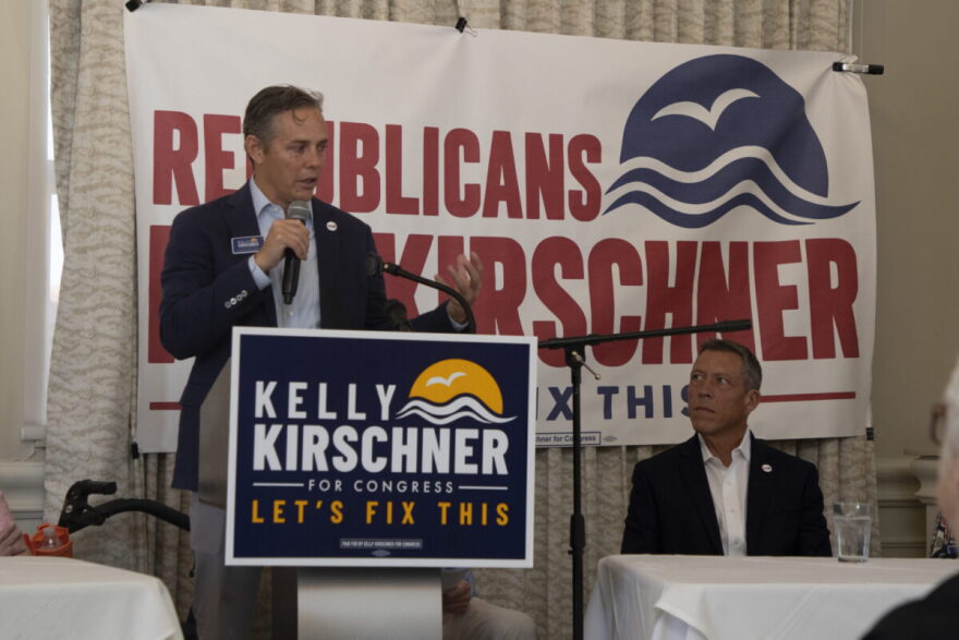 A man with dark hair stands on a small stae behind a sign that says kelly kirschner for congress "let's fix this." He's speaking into a mic.