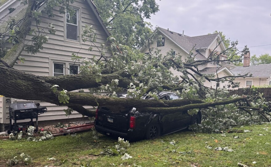 Broken tall tree fallen onto a car parked in a yard next to a house