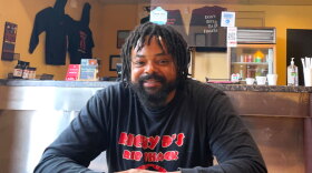 A man sits at a table in a restaurant. He's wearing a black long sleeve shirt with red lettering that reads "Ricky D's Rib Shack. He sits in front of a counter.