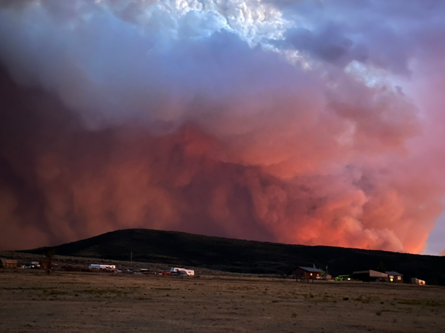 Thick clouds of red and gray wildfire smoke loom over a hill in a rural area.