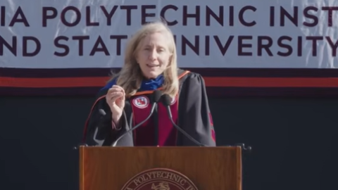A woman with blonde hair stands at a podium, wearing a black robe with a maroon sash. 