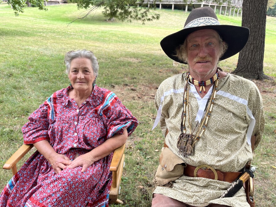 A Cherokee couple sit next to each other outside; the woman wearing a red Tear Dress and the man is wearing a hat and tan linen shirt with tribal jewelry.