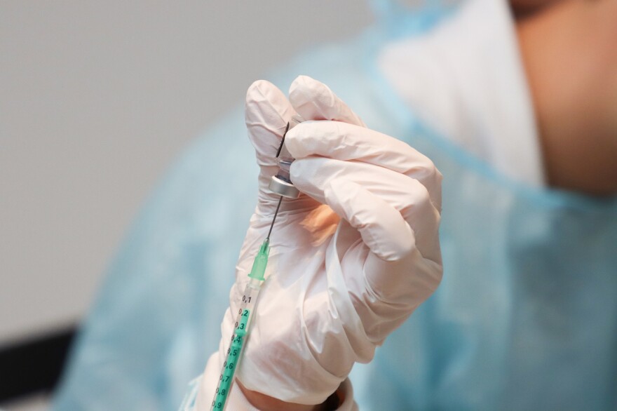 Closeup of a gloved hand holding a small glass vaccine bottle while contents are being extracted with a synringe.